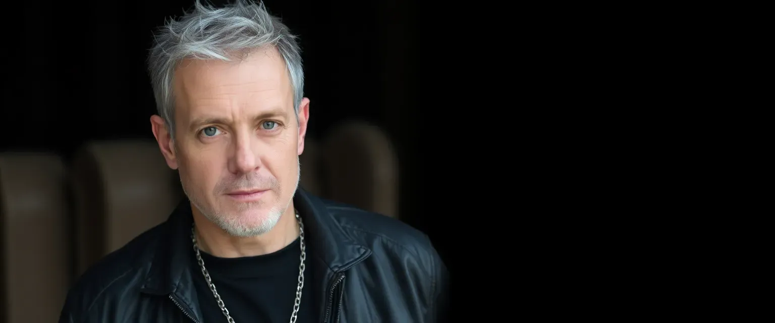 Man in his 40s, graying short hair, piercing gray eyes, wearing a leather jacket, black t-shirt, dark jeans, silver chain.